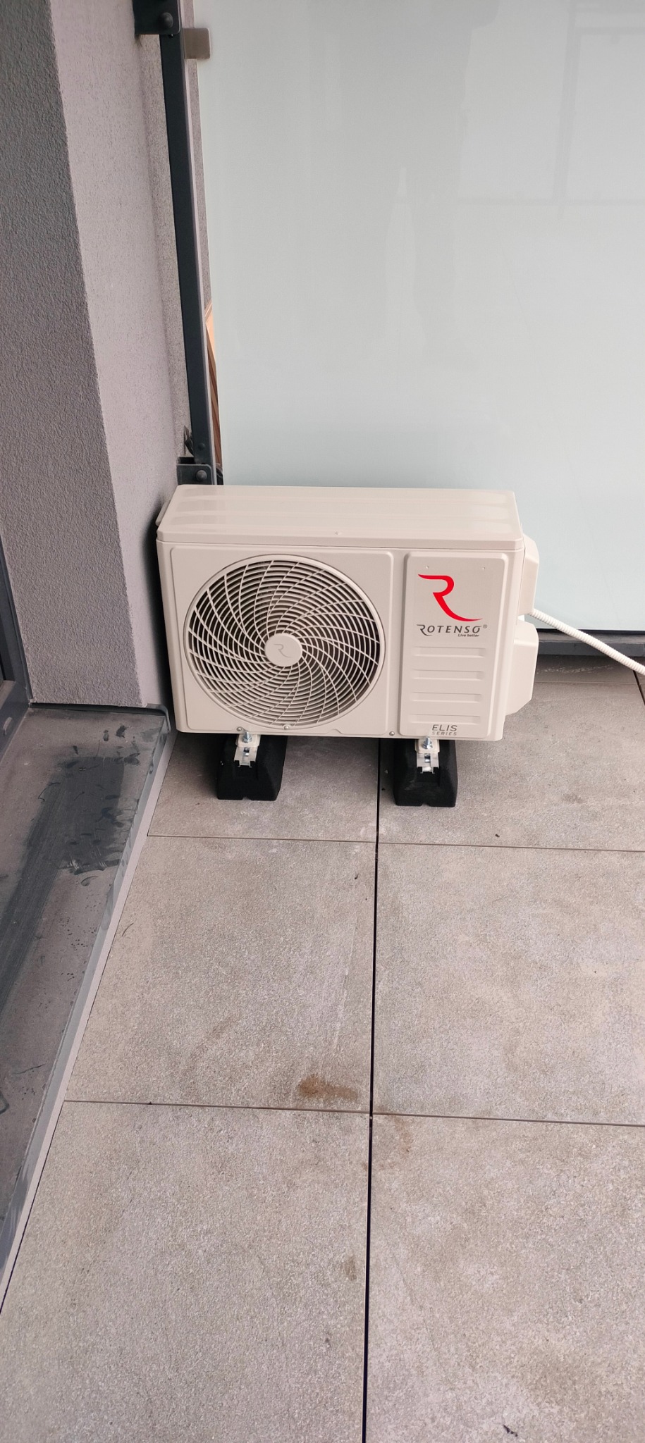 Kompaktowa jednostka zewnętrzna klimatyzacji Rotenso Elis Series ustawiona na czarnych podkładkach antywibracyjnych na balkonie z jasnych płytek.