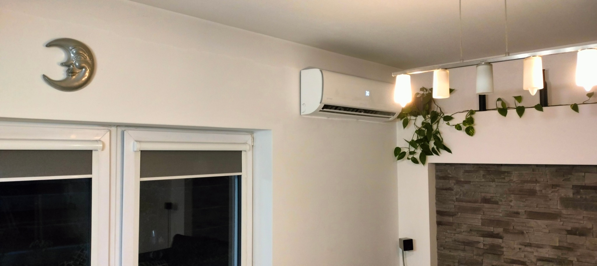 Biały klimatyzator ścienny z wyświetlaczem temperatury, zamontowany w jasnym pokoju z oknami, dekoracją w kształcie księżyca i roślinami zwisającymi z lampy.