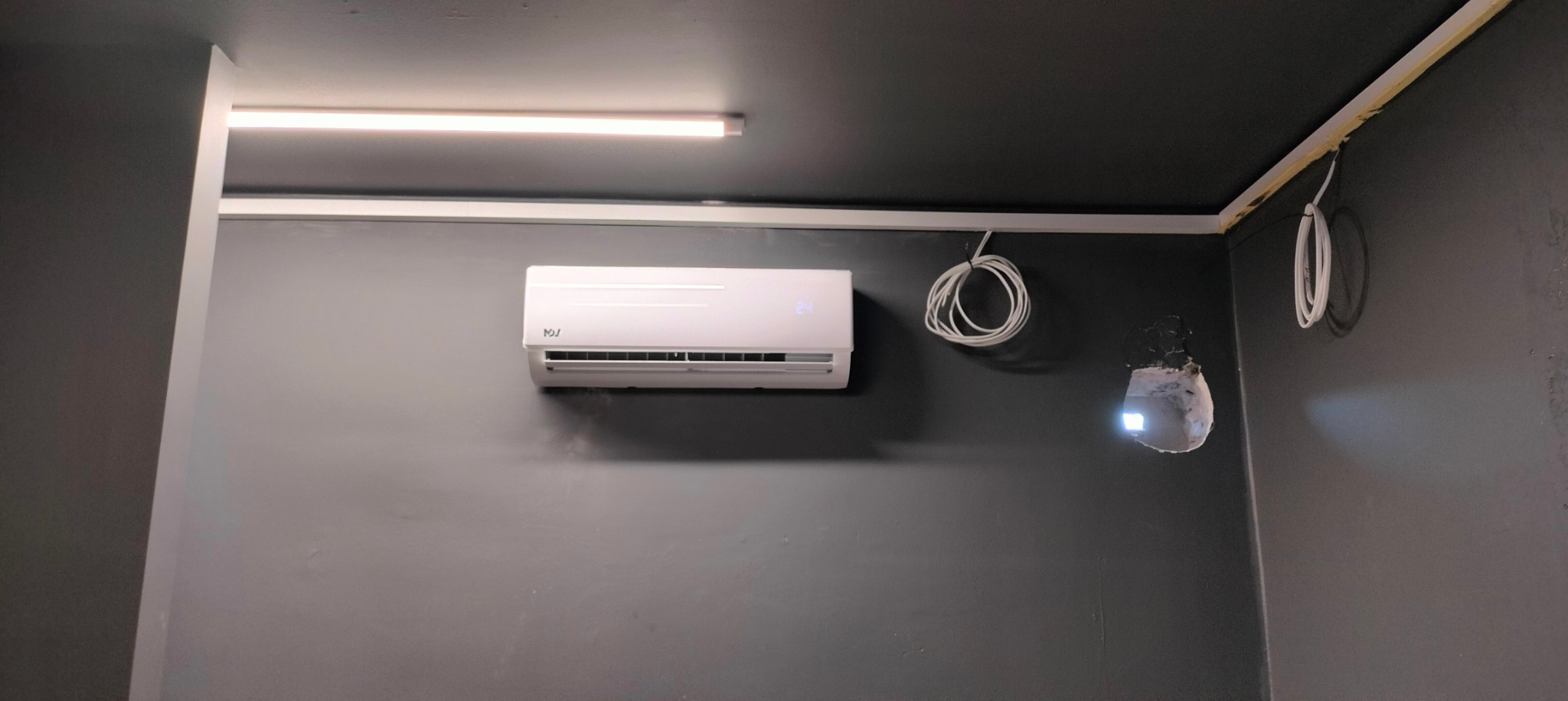 Biały klimatyzator ścienny na szarej ścianie, widoczne kable i otwór w ścianie po lewej stronie, na suficie widoczna ledowa lampa.