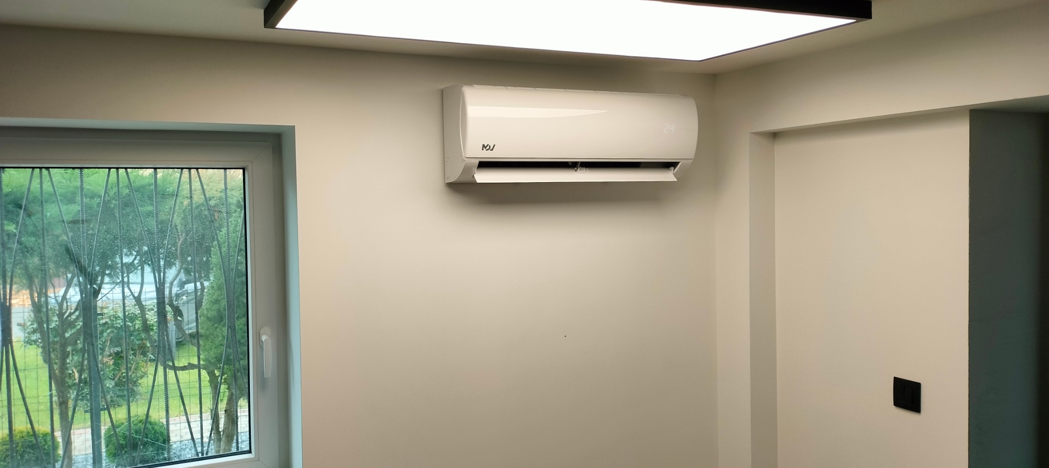 Biała klimatyzacja ścienna z wyświetlaczem temperatury 24, zamontowana w jasnym pokoju z oknem z ozdobną kratą, widok na zieleń na zewnątrz.