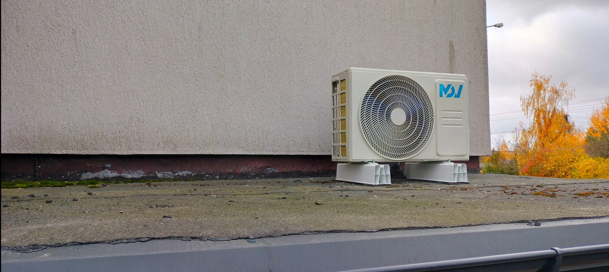 Zewnętrzna jednostka klimatyzacji zamontowana na dachu budynku, widoczna na tle ściany i jesiennych drzew.
