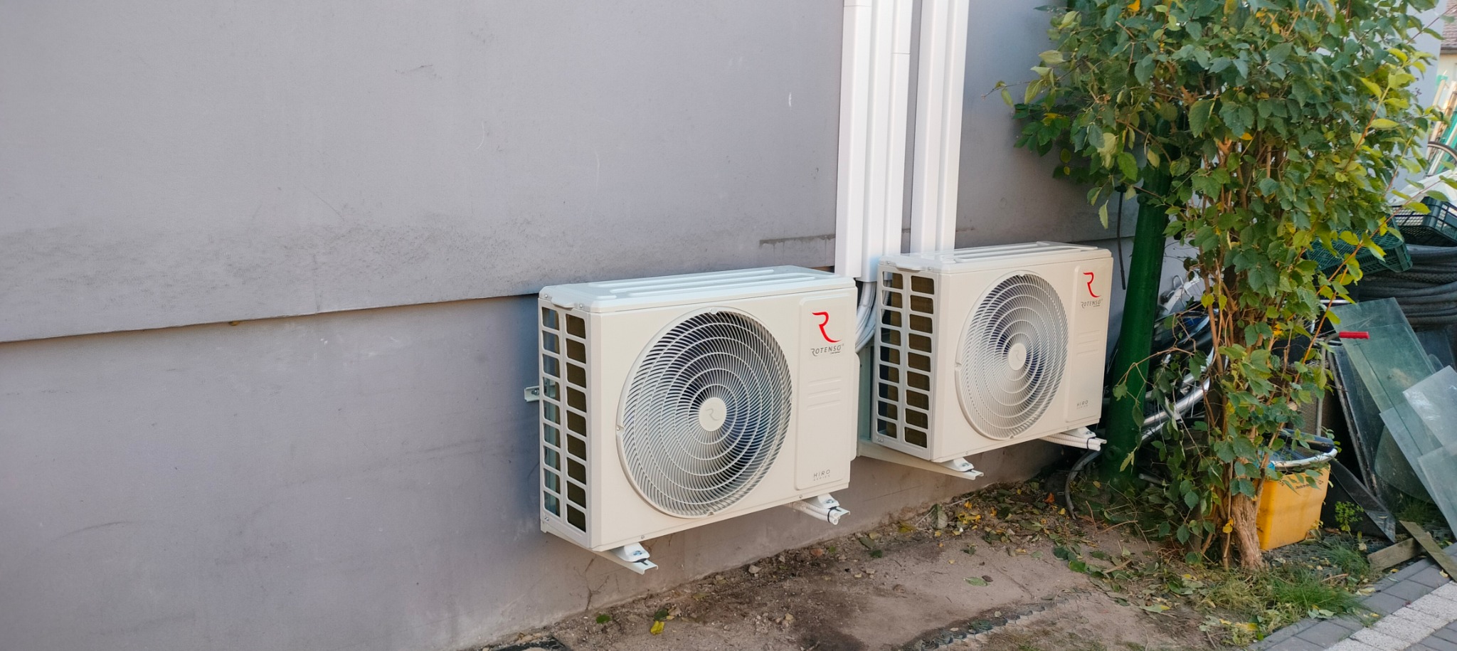 Dwa białe klimatyzatory Rotenso Hiro zamontowane na szarej ścianie budynku, widoczne rury instalacyjne i fragment zieleni w tle.
