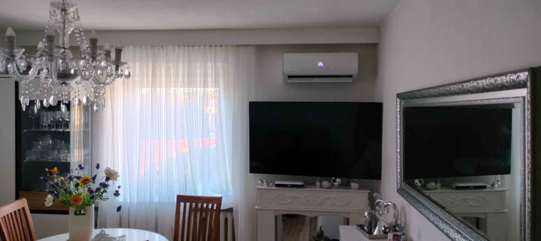 Biały klimatyzator ścienny z wyświetlaczem temperatury 26 stopni Celsjusza, zamontowany nad telewizorem w salonie z kryształowym żyrandolem i dekoracyjnym kominkiem.