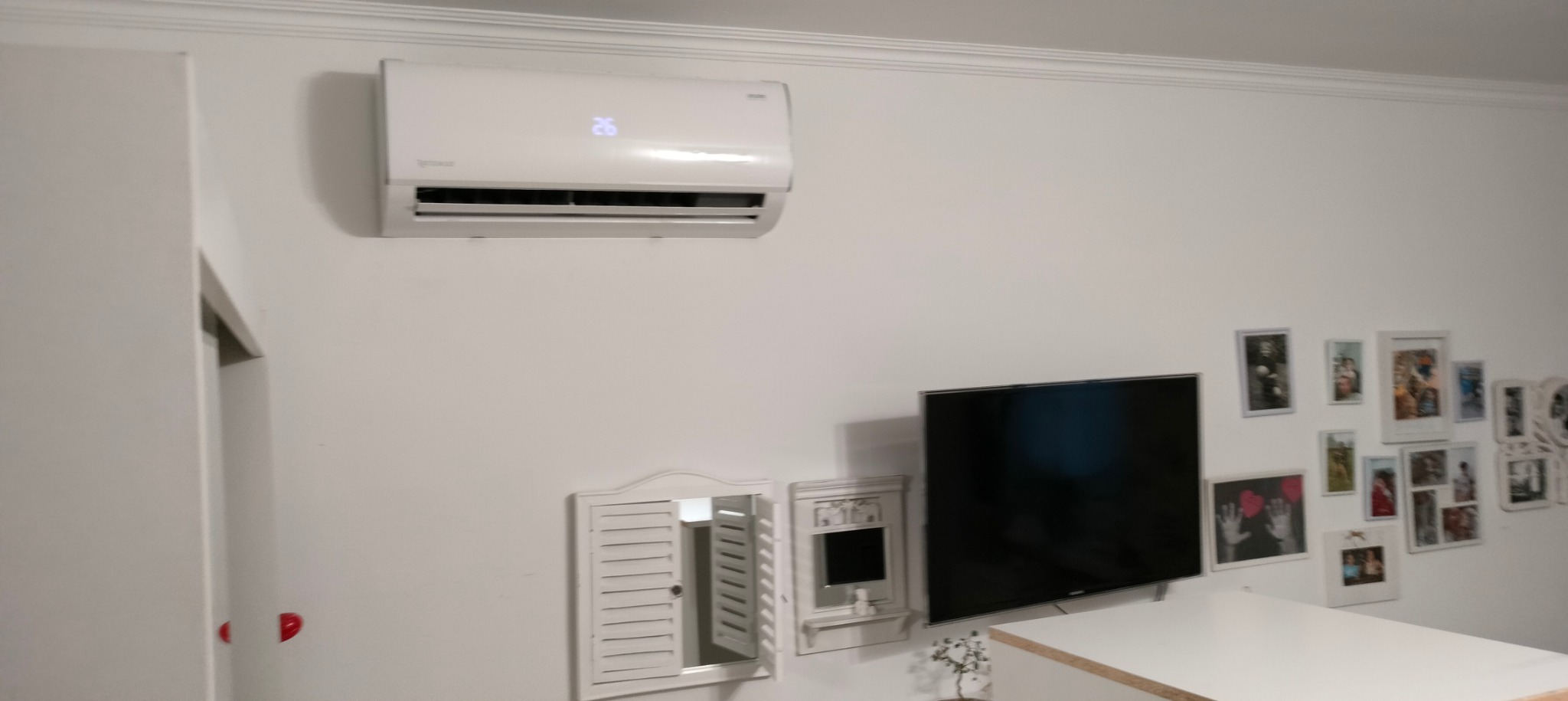 Biały klimatyzator ścienny z wyświetlaczem temperatury 26 stopni Celsjusza, zamontowany w pokoju z dekoracjami ściennymi i telewizorem.