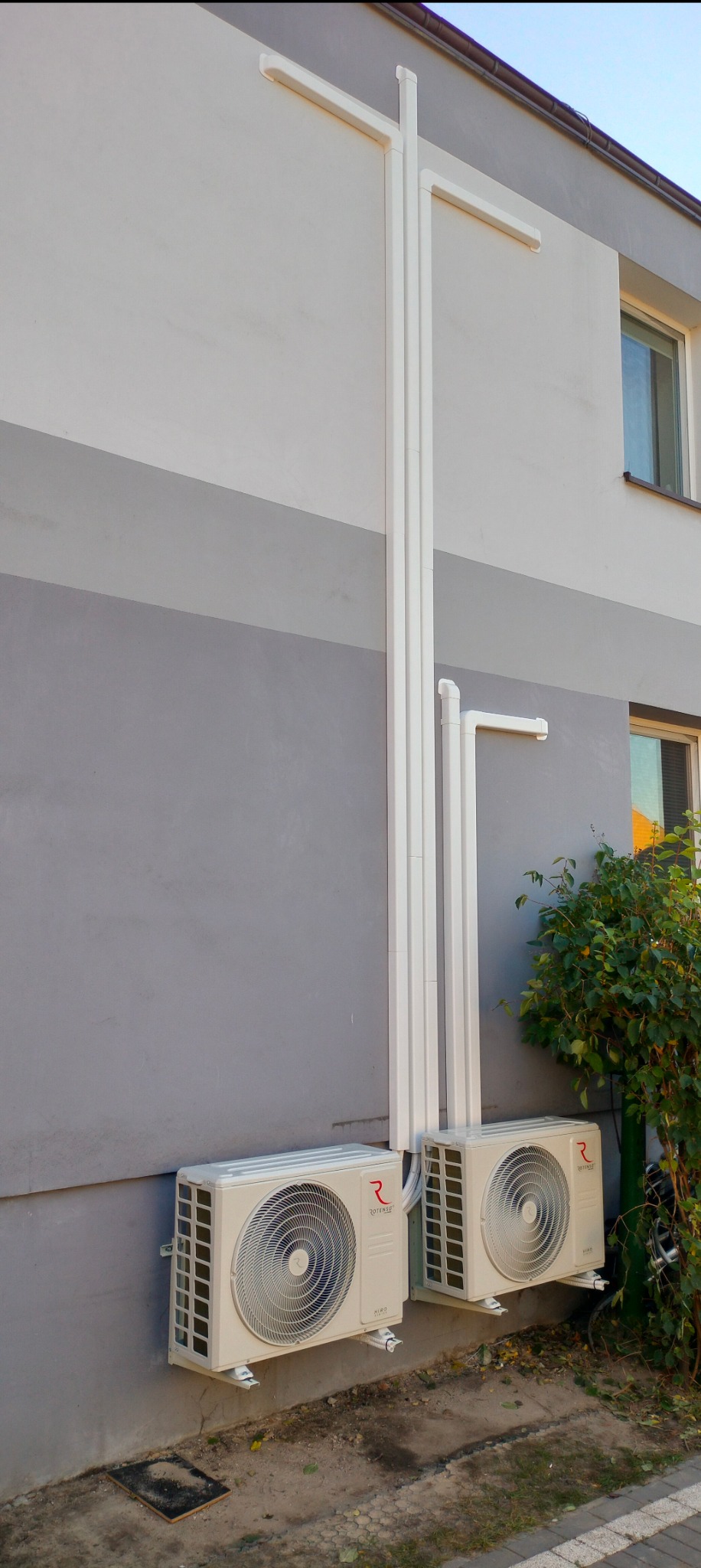 Dwa klimatyzatory zamontowane na szarej ścianie budynku, z widocznymi białymi rurami prowadzącymi w górę, oraz krzewem po prawej stronie.