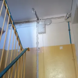 Szara skrzynka elektryczna z widocznymi przewodami na klatce schodowej w bloku. U dołu ściana pomalowana na żółto, u góry na biało. Widoczne schody z niebieską barierką.