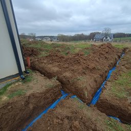 Usługi Elektryczne i Pomiarowe Aleksandra Ozga - Wykopane rowy w ziemi, wyłożone niebieską folią, prawdopodobnie pod instalacje. W tle widoczne domy jednorodzinne i pochmurne niebo.
