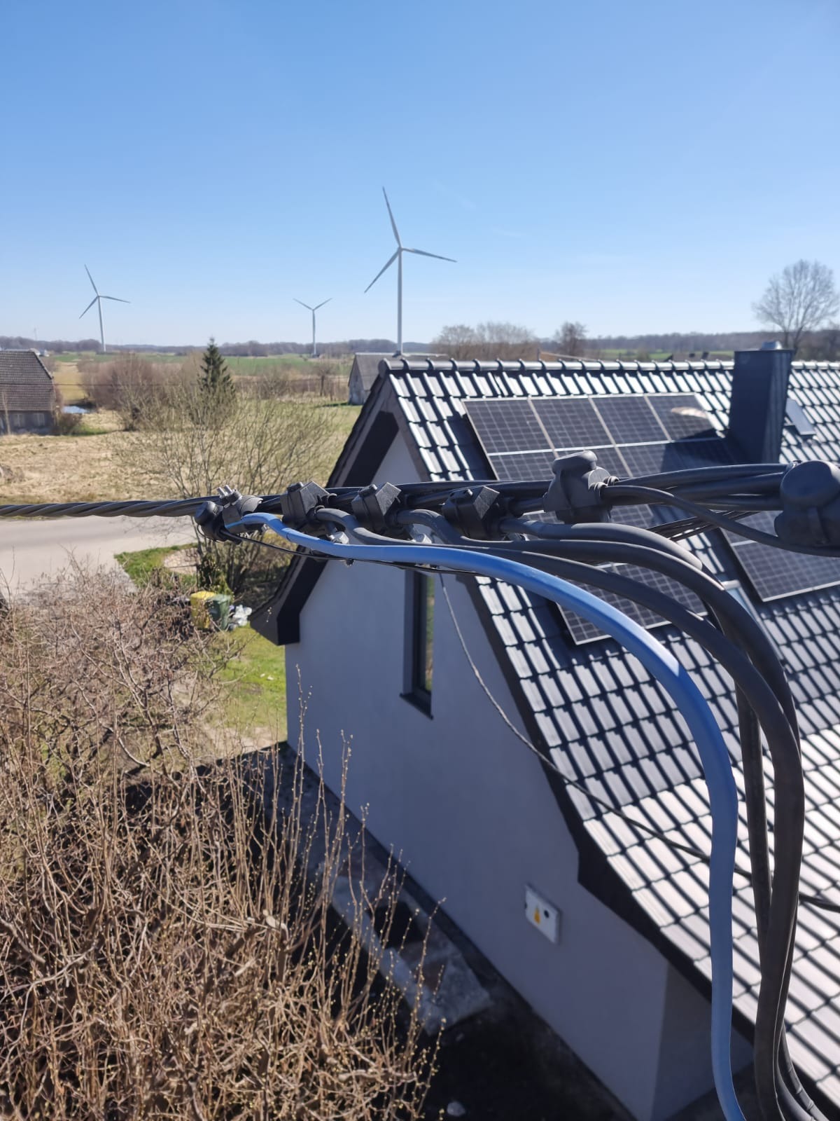 Przyłącze kablowe do domu z panelami solarnymi na dachu. W tle widać turbiny wiatrowe. Kable poprowadzone są nad budynkiem.