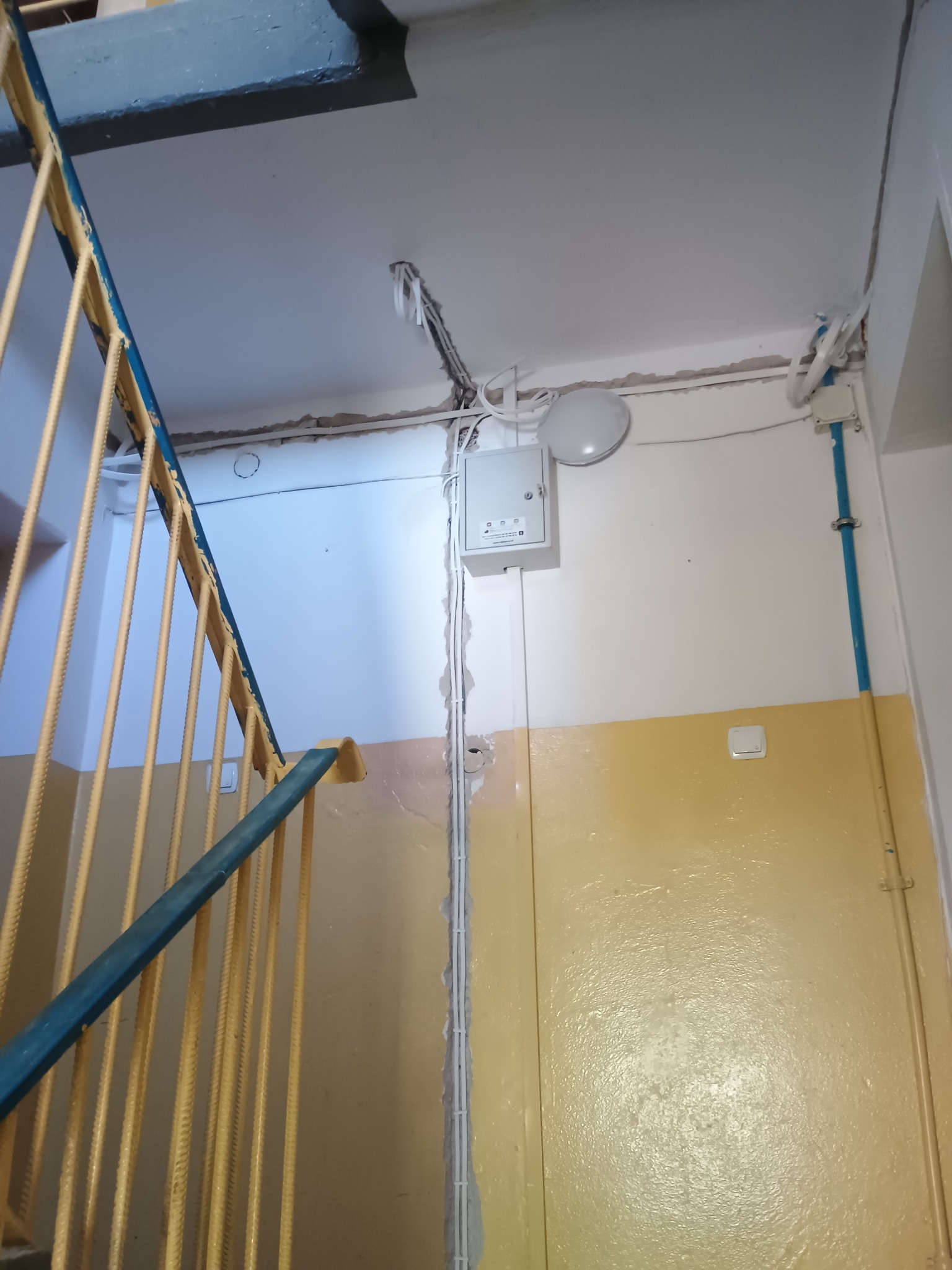 Szara skrzynka elektryczna z widocznymi przewodami na klatce schodowej w bloku. U dołu ściana pomalowana na żółto, u góry na biało. Widoczne schody z niebieską barierką.