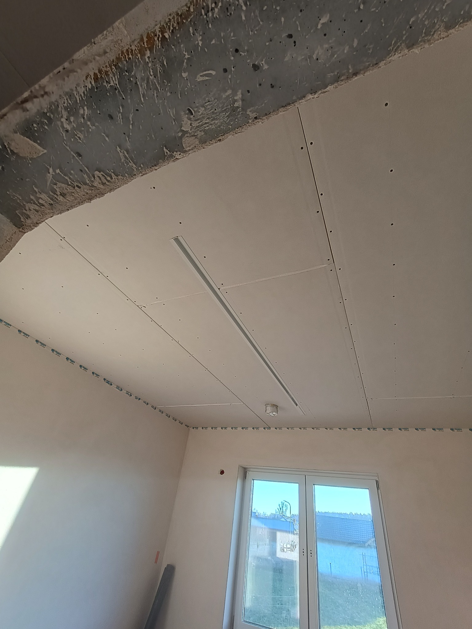 Zamontowany sufit podwieszany z płyt gipsowo-kartonowych, widoczna listwa oświetleniowa i okno z widokiem na zewnątrz. Prace wykończeniowe w nowym budynku.