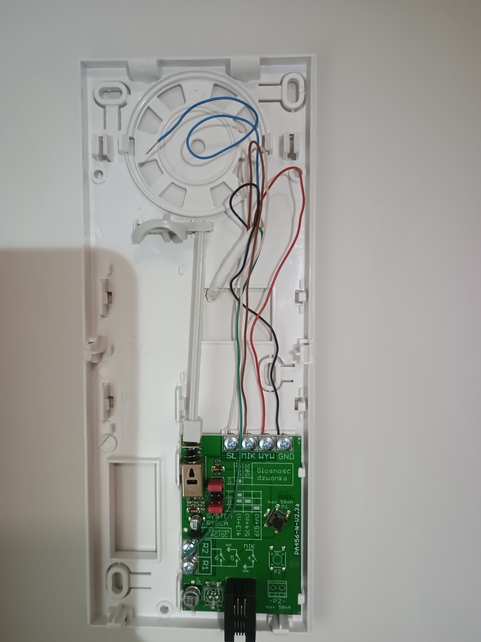 Otwarta obudowa domofonu z widoczną płytką drukowaną, przewodami i elementami montażowymi na białym tle. Widoczne ślady po montażu.