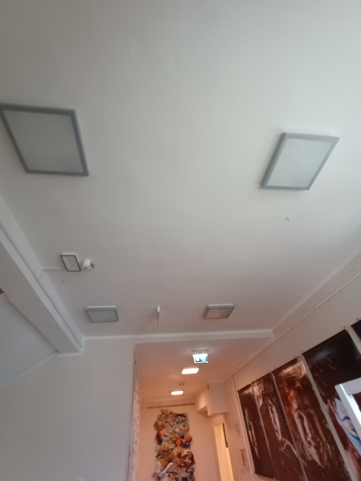 Biały sufit z zamontowanymi, kwadratowymi lampami LED w szarych ramkach. Dodatkowo widoczna kamera przemysłowa i oświetlenie awaryjne.