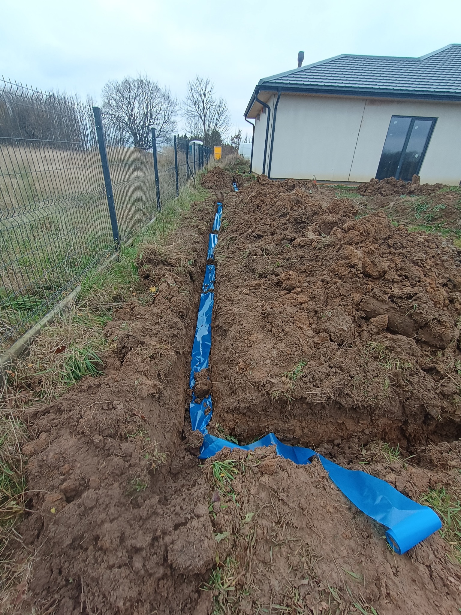 Wykop w ziemi z niebieską folią zabezpieczającą, prawdopodobnie pod instalację elektryczną, widok z góry, w tle dom jednorodzinny i ogrodzenie.