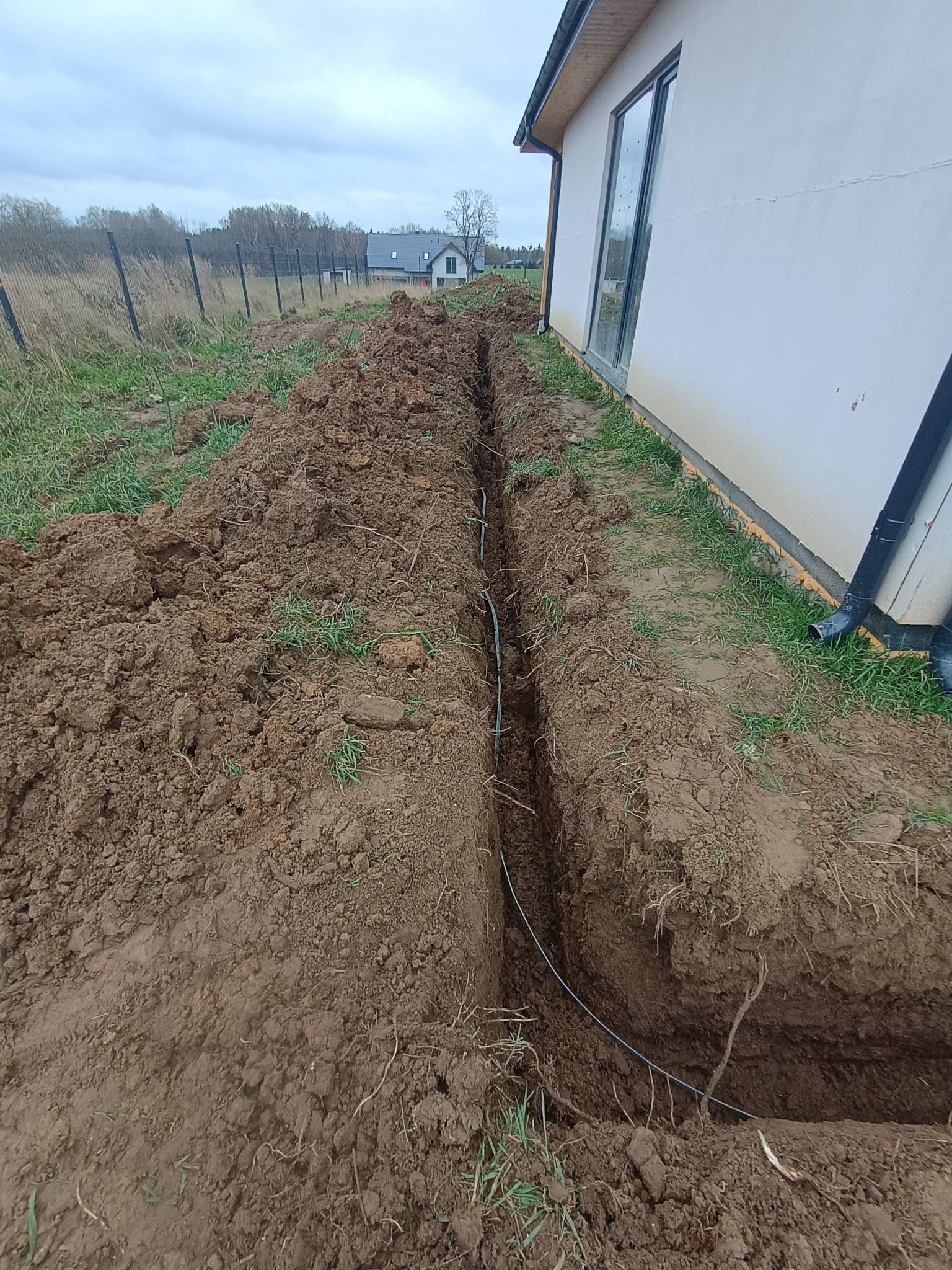 Wykopany rów w ziemi, w którym ułożony jest kabel. Widoczny fragment budynku z oknem i rynną. Trawa i ogrodzenie w tle. Pochmurne niebo.