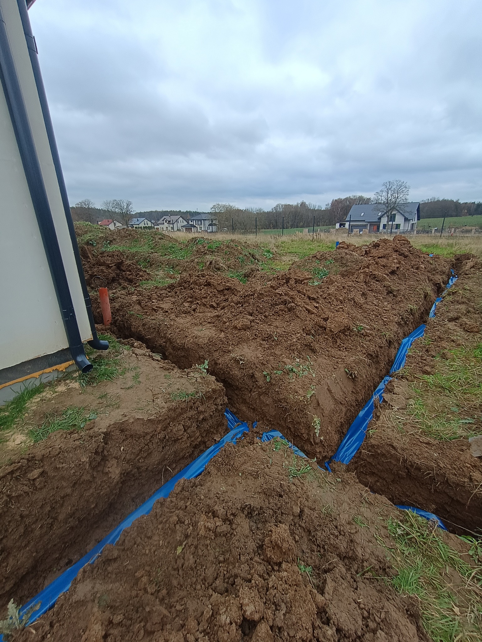 Wykopane rowy w ziemi, wyłożone niebieską folią, prawdopodobnie pod instalacje. W tle widoczne domy jednorodzinne i pochmurne niebo.