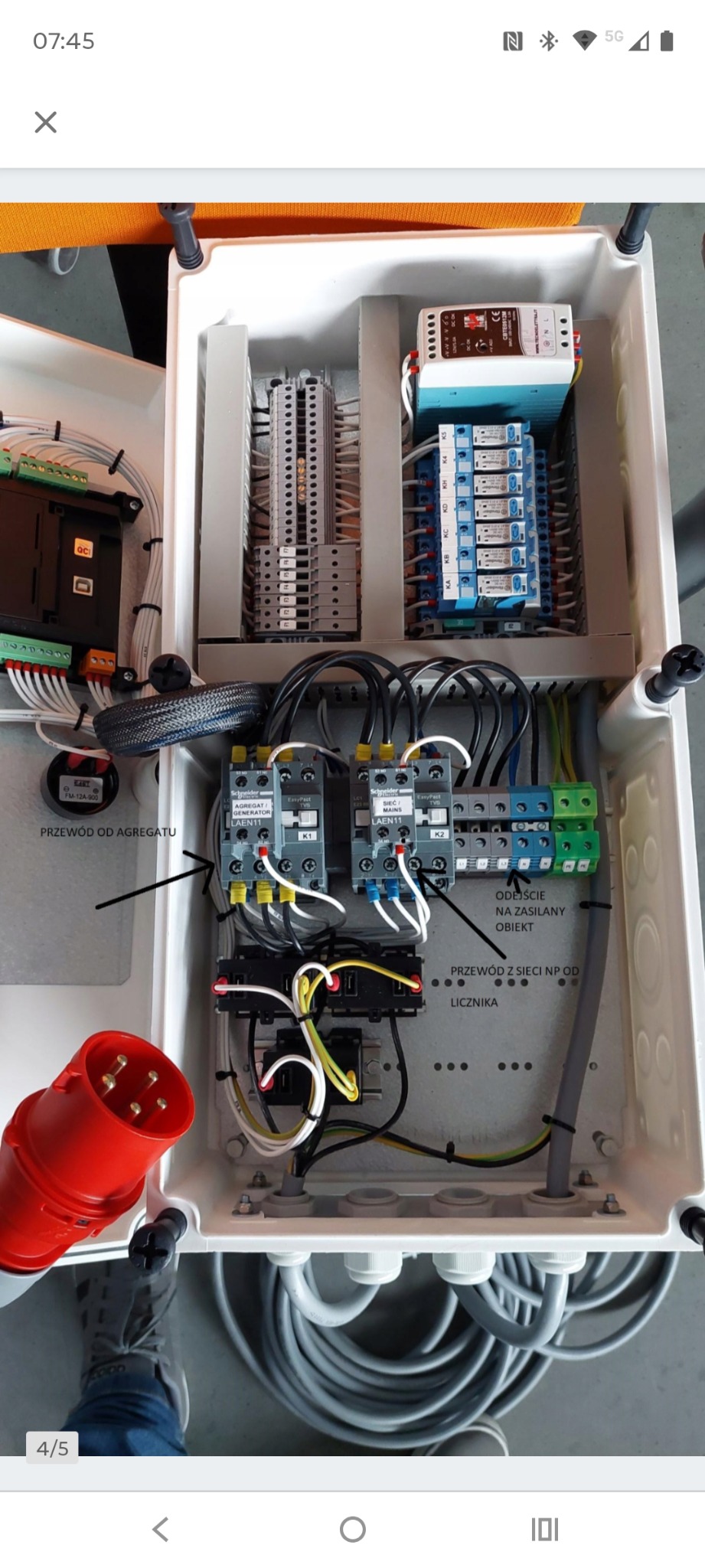 Otwarta skrzynka elektryczna z widocznymi elementami instalacji: styczniki, przekaźniki, listwy zaciskowe. Widoczne przewody i oznaczenia. Czerwona wtyczka na dole.