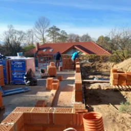 Wznoszenie ścian z ceramicznych pustaków na budowie domu jednorodzinnego, widoczne palety z materiałem Leier Plan, trzech pracowników przy pracy, w tle dom z czerwoną dachówką i błękitne niebo.