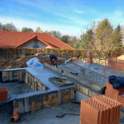 Zalewanie fundamentu betonem w szalunku drewnianym, widoczne zbrojenie, w tle budynek z czerwoną dachówką i błękitne niebo.