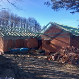 BMV MS - Wznoszenie ścian z czerwonej cegły domu jednorodzinnego z widocznym drewnianym szkieletem dachu i stertą cegieł na błotnistym terenie budowy w słoneczny dzień.