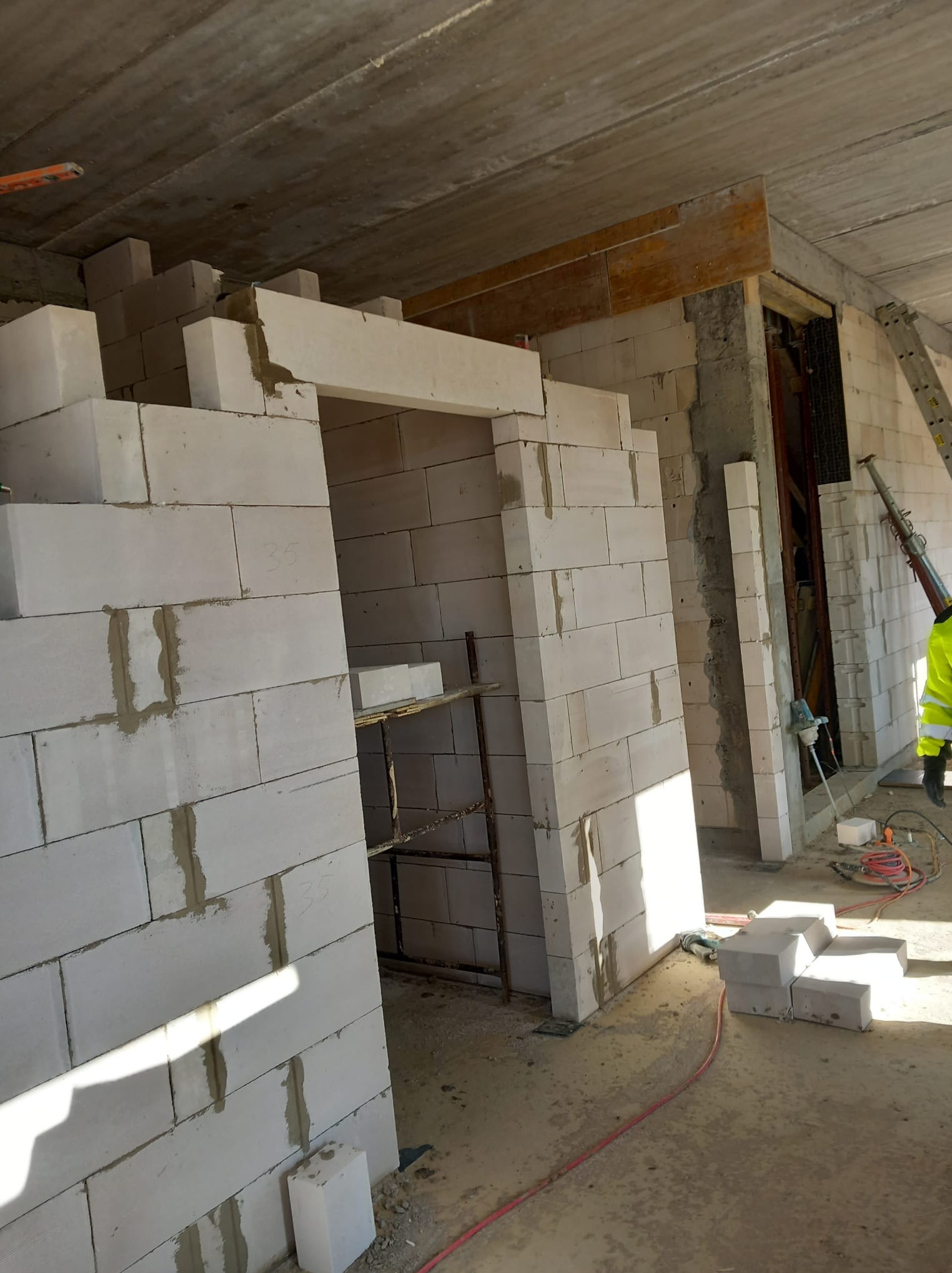 Wznoszenie ścian z białych bloczków w surowym stanie, widoczny szary strop i fragment rusztowania, ślady zaprawy murarskiej między bloczkami, pracownik w odblaskowej kamizelce w tle.