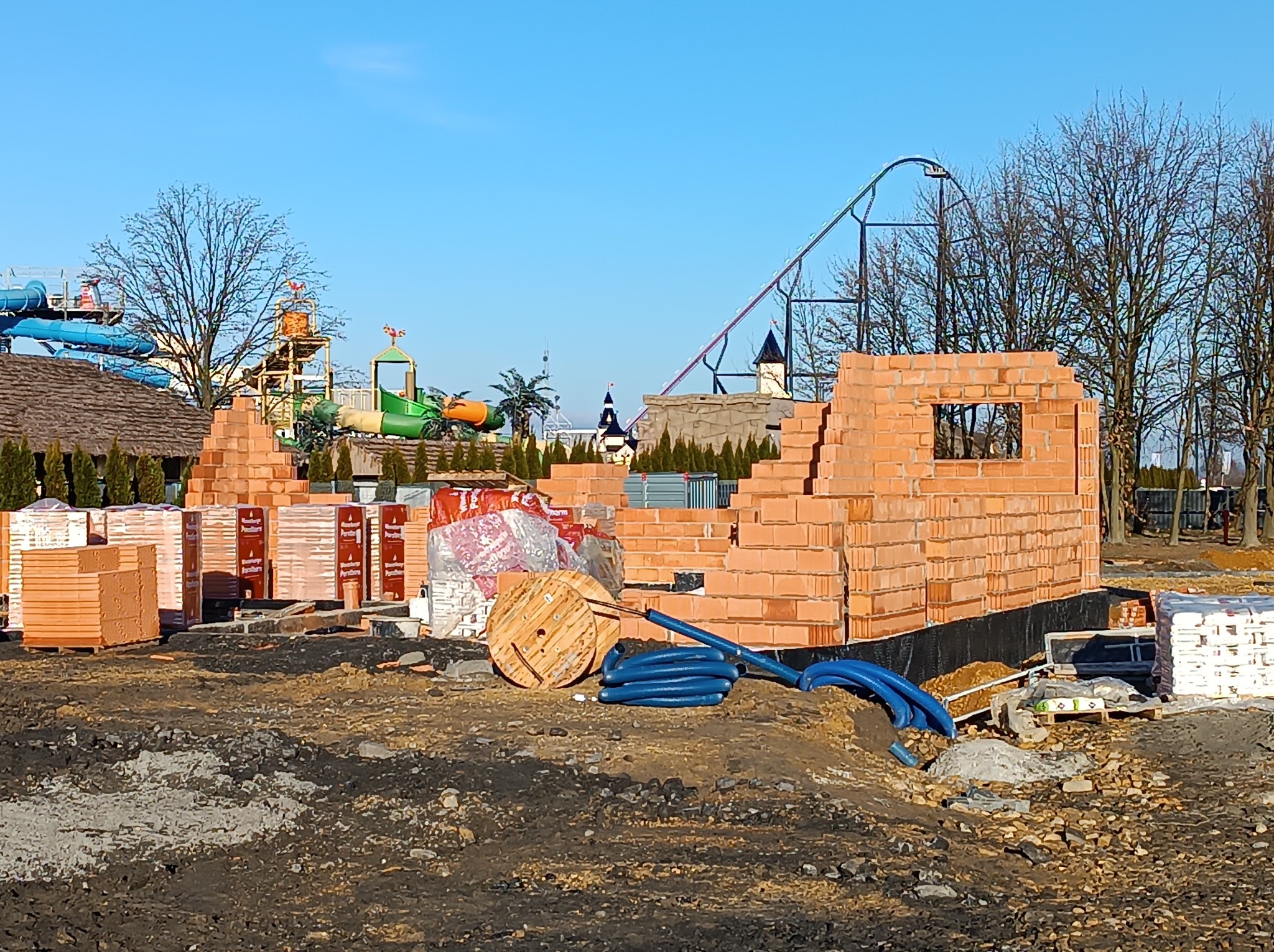 Wznoszone ściany z cegły ceramicznej na placu budowy z widokiem na park rozrywki z rollercoasterem w tle, niebieskie rury na pierwszym planie.