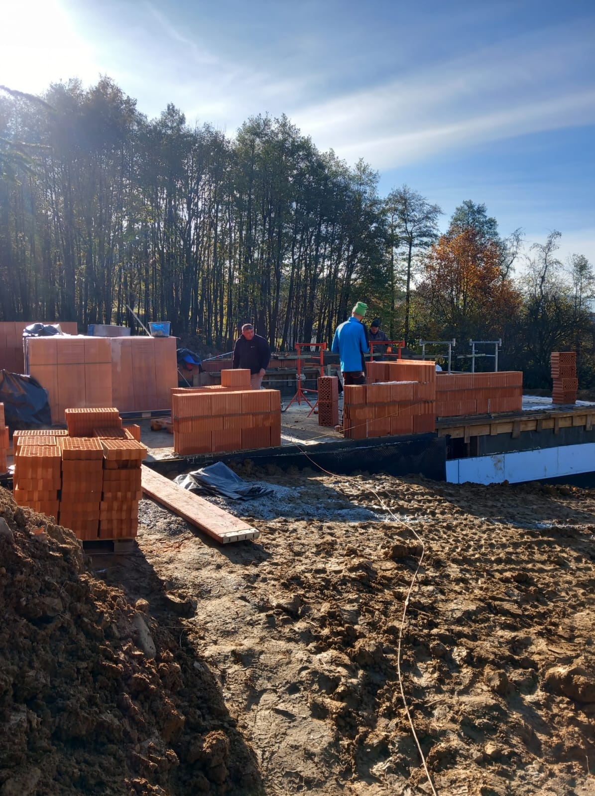 Wczesny etap budowy domu: fundamenty przygotowane, wznoszone ściany z czerwonej cegły ceramicznej, widoczni dwaj pracownicy w trakcie murowania, w tle drzewa i błękitne niebo.