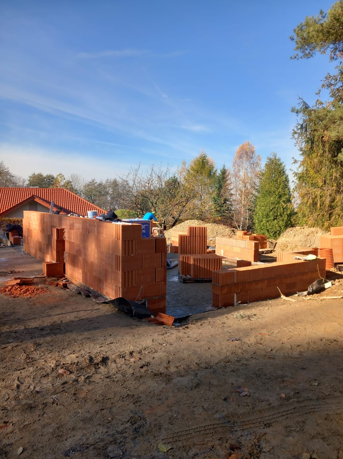 Wznoszenie ścian parterowego domu z czerwonej ceramiki poryzowanej, widoczni pracownicy w trakcie murowania, palety z bloczkami obok, jesienne drzewa w tle.