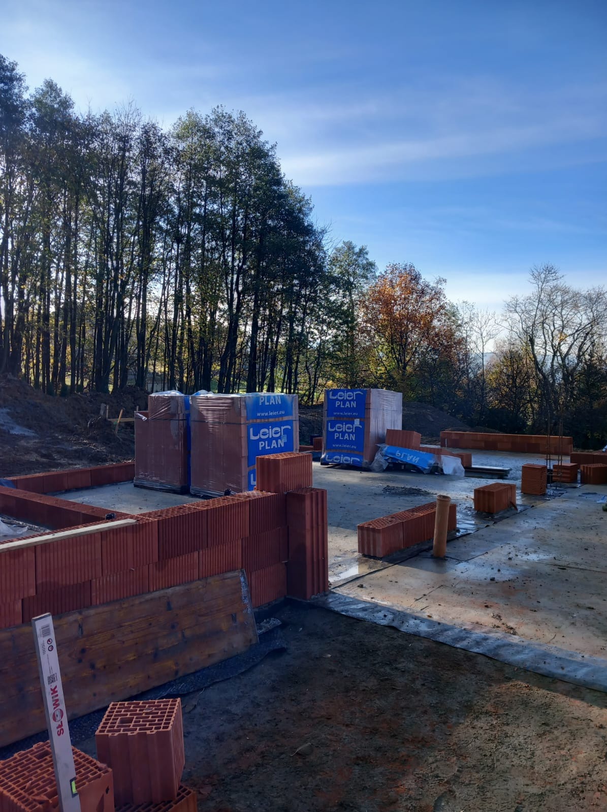 Wczesny etap budowy domu: widoczne fundamenty z czerwonej cegły ceramicznej, palety z pustakami Leier Plan w niebieskiej folii, poziomica przy murze, drzewa w tle pod błękitnym niebem.