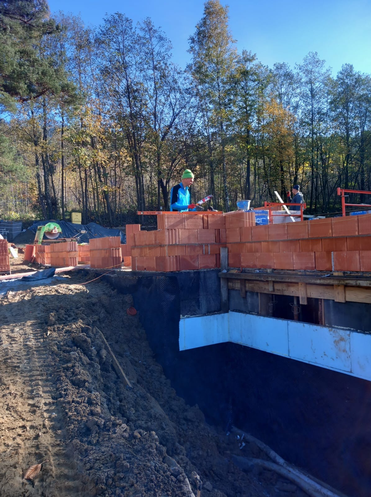 Budowa domu: murarz w zielonej czapce łączy pustaki ceramiczne na fundamencie z hydroizolacją, widoczne drzewa w tle.