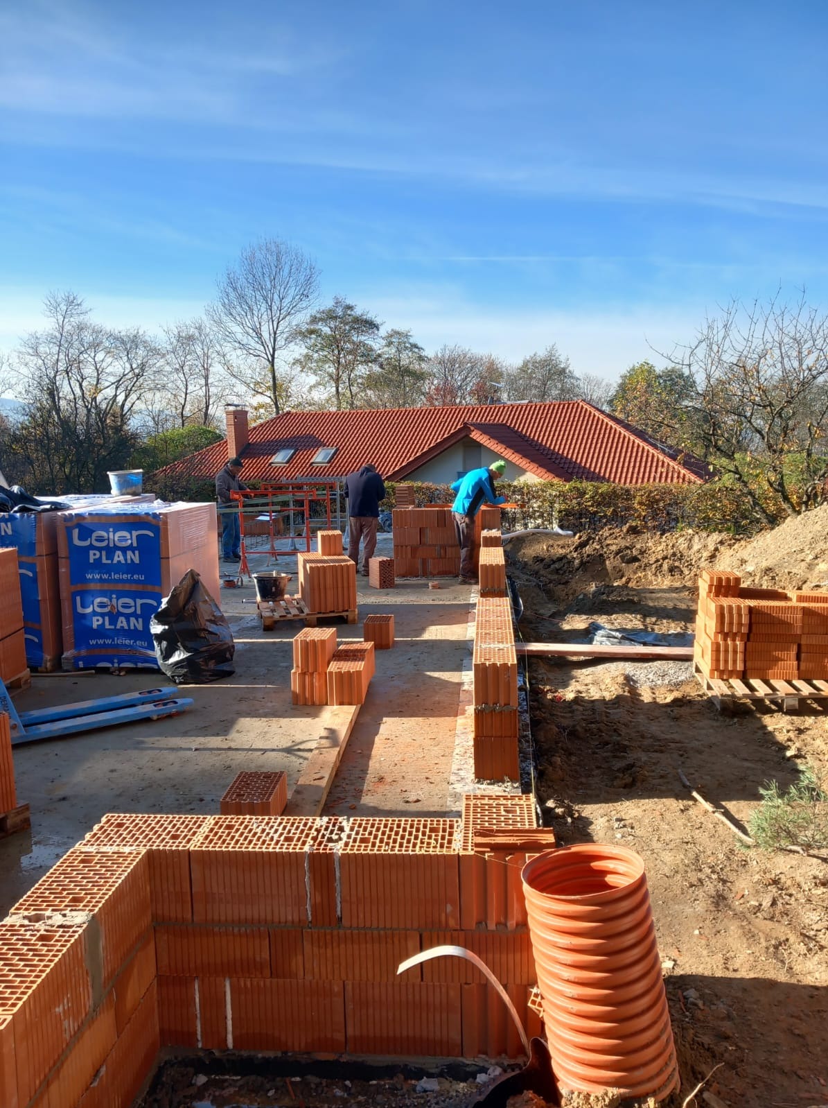Wznoszenie ścian z ceramicznych pustaków na budowie domu jednorodzinnego, widoczne palety z materiałem Leier Plan, trzech pracowników przy pracy, w tle dom z czerwoną dachówką i błękitne niebo.