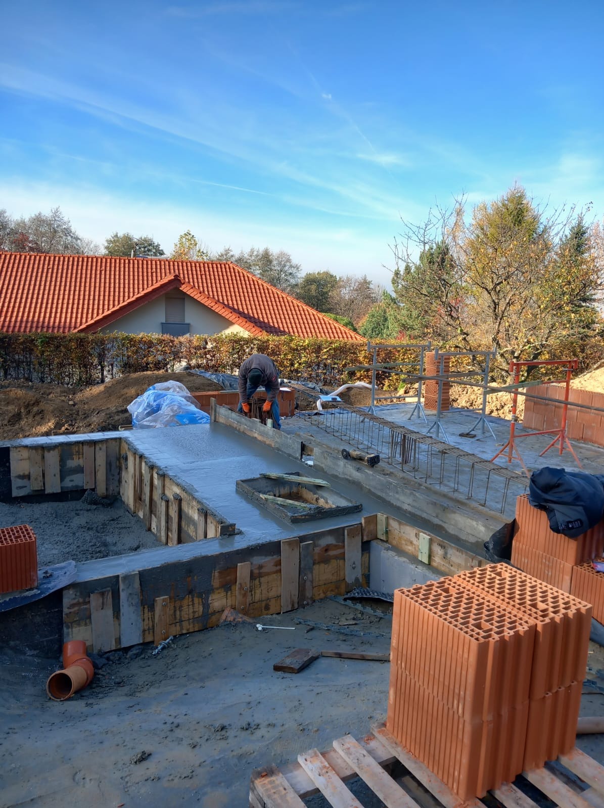 Zalewanie fundamentu betonem w szalunku drewnianym, widoczne zbrojenie, w tle budynek z czerwoną dachówką i błękitne niebo.