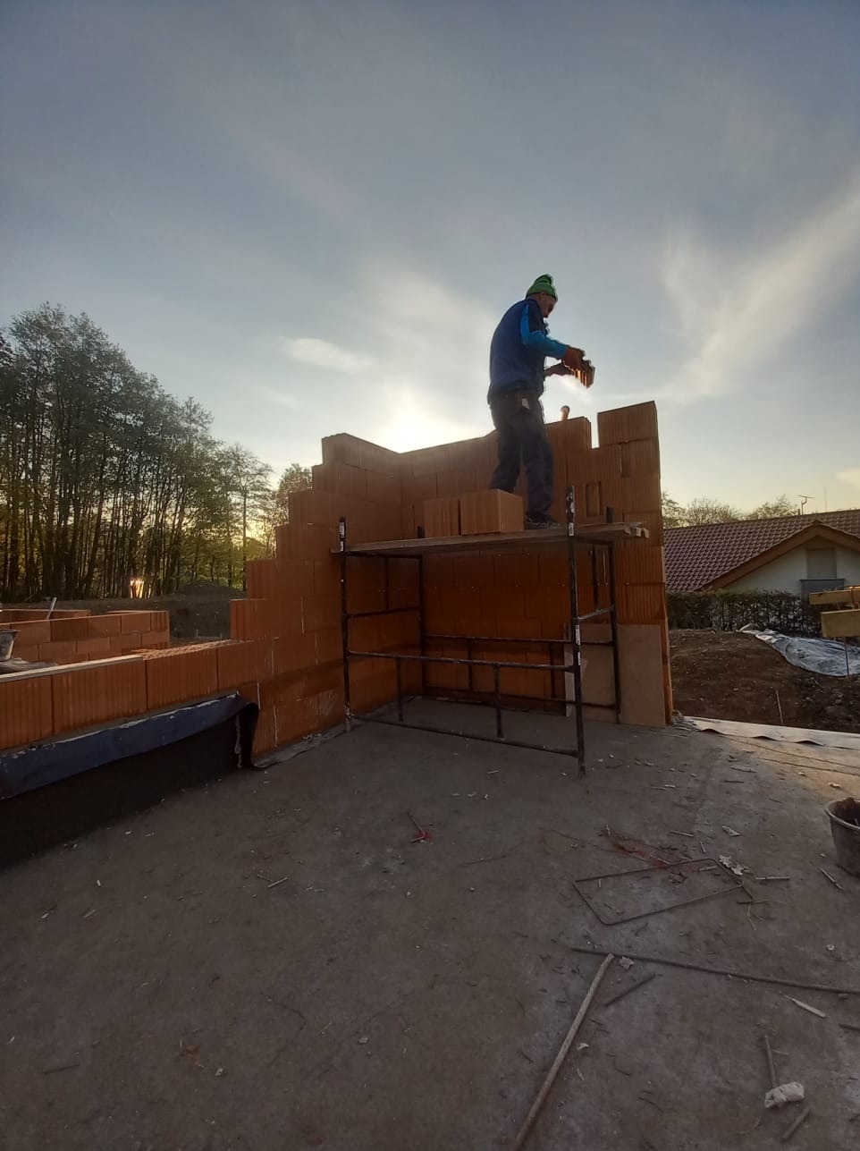 Murarz na rusztowaniu układa cegły na wznoszonej ścianie domu, widoczne drzewa w tle.