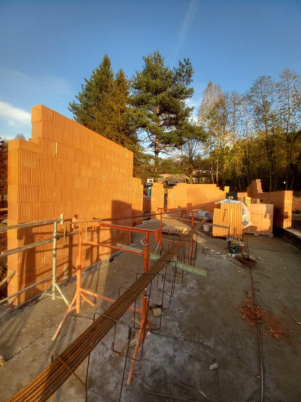 Wznoszenie ścian z pustaków ceramicznych na placu budowy, widoczne rusztowanie, zbrojenie i drzewa w tle pod błękitnym niebem.