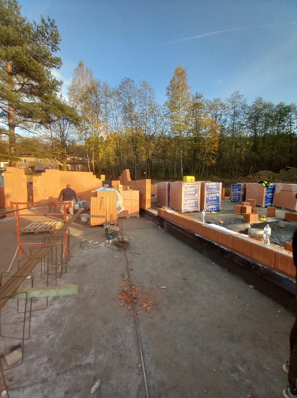 Wznoszenie ścian domu z bloczków ceramicznych, widoczne palety z materiałem budowlanym, zbrojenie fundamentów i pracujący murarz w tle, jesienne drzewa i błękitne niebo.