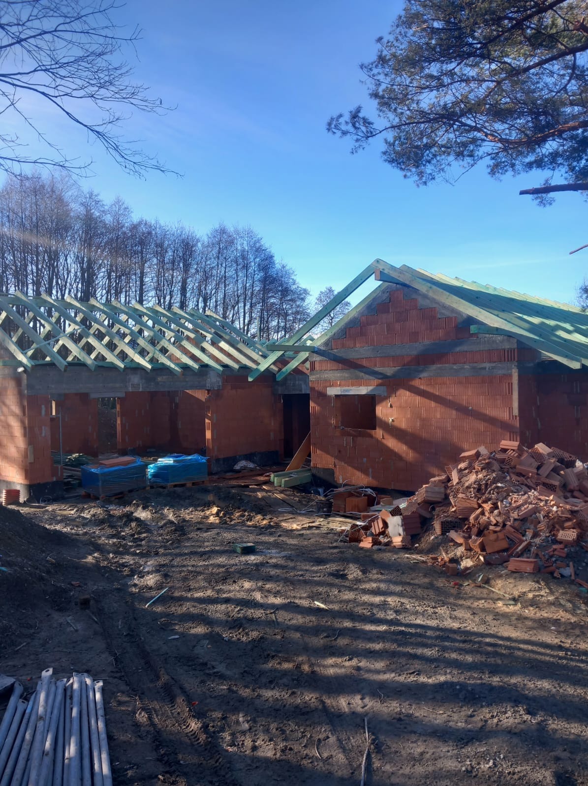Wznoszenie ścian z czerwonej cegły domu jednorodzinnego z widocznym drewnianym szkieletem dachu i stertą cegieł na błotnistym terenie budowy w słoneczny dzień.