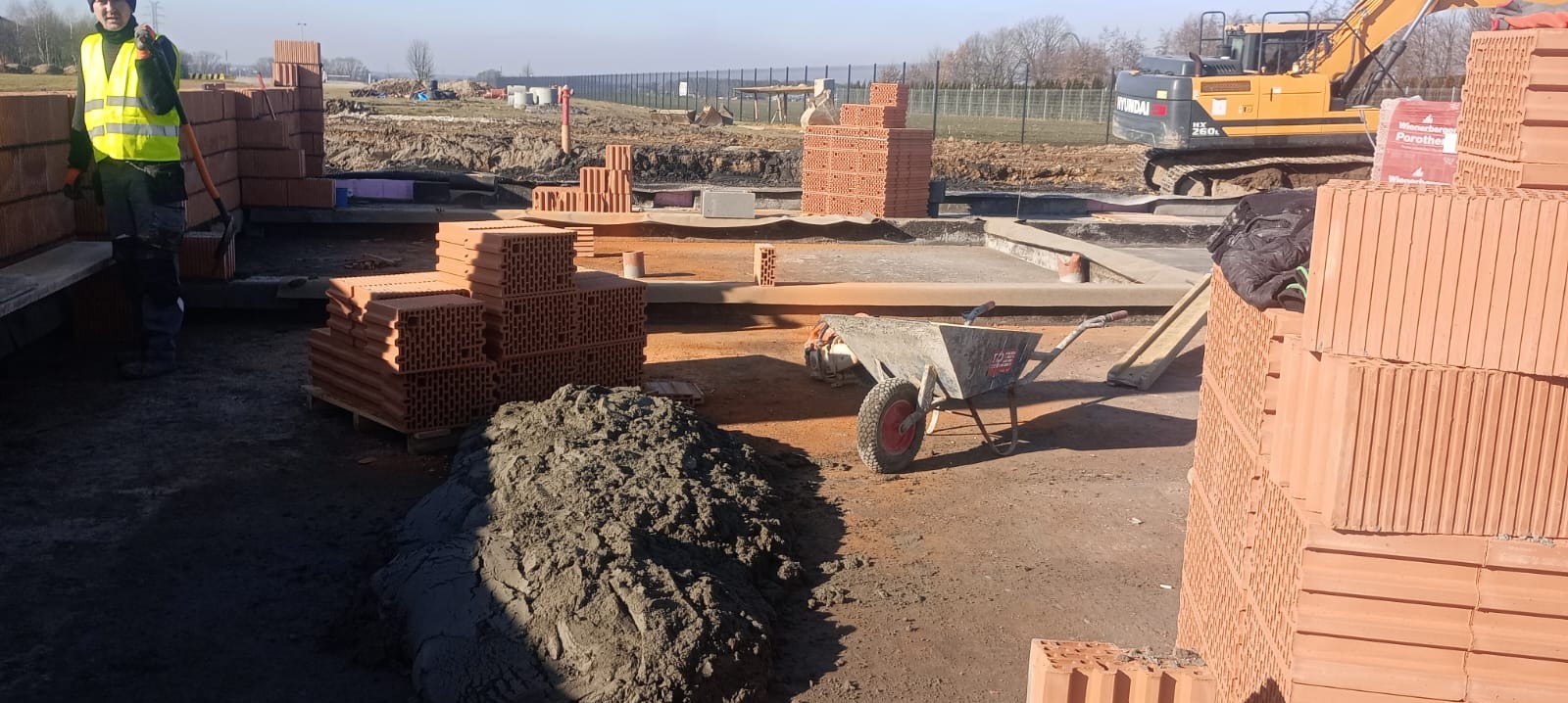 Budowa fundamentów: widok na wykop z betonową wylewką, stosy cegieł Porotherm, taczkę i koparkę Hyundai w tle, pracownik w kamizelce odblaskowej przy częściowo wymurowanej ścianie.