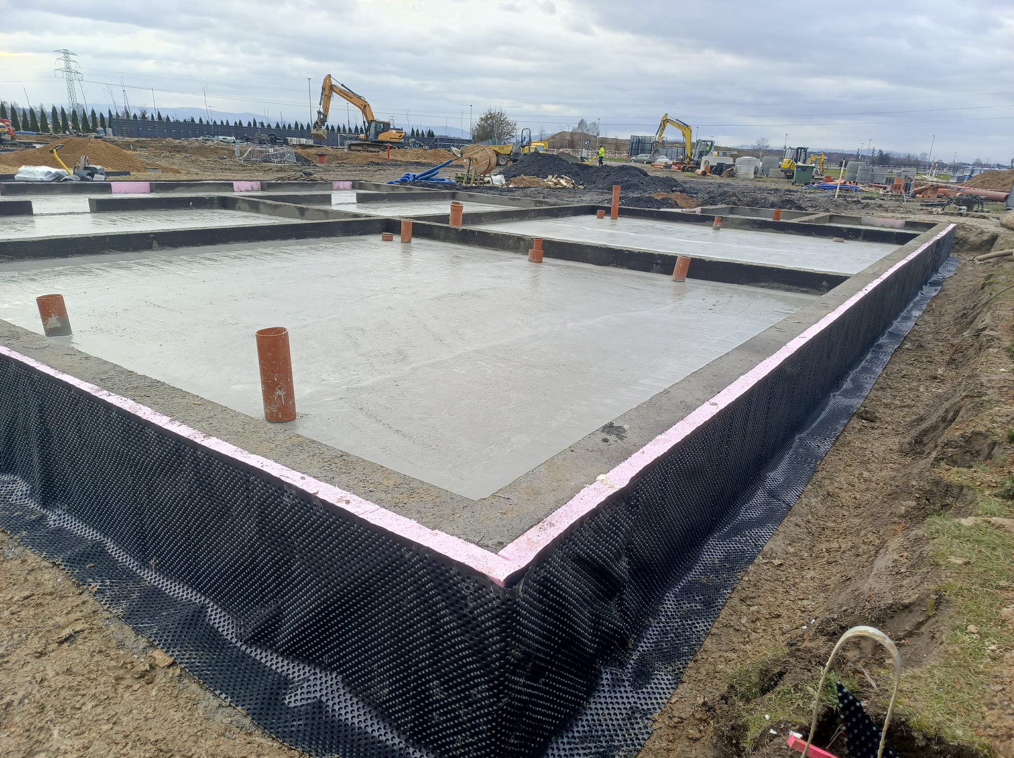 Wylany fundament z hydroizolacją pionową i rurami kanalizacyjnymi wystającymi ponad powierzchnię, z koparkami w tle na placu budowy.