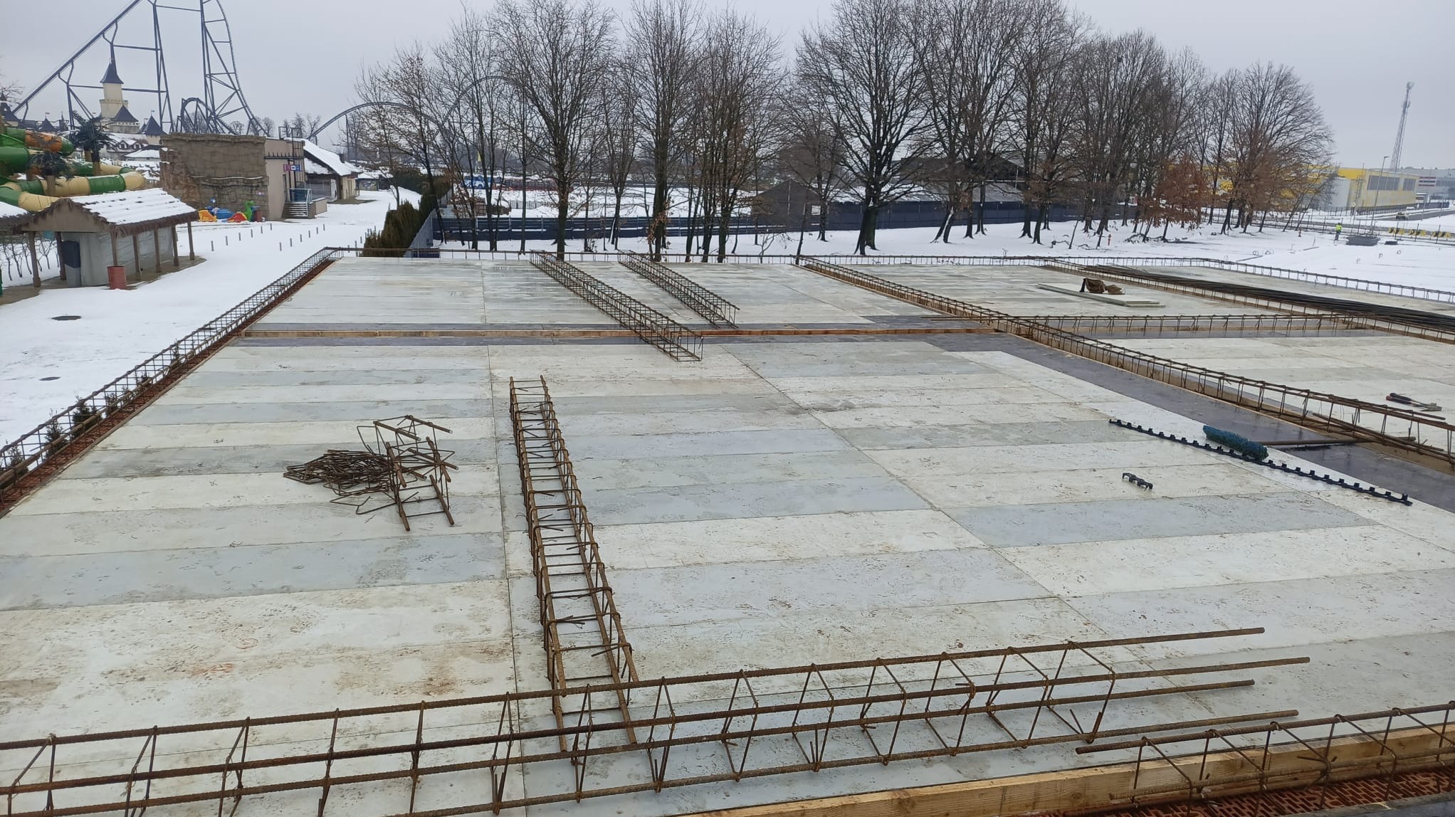 Przygotowany szalunek i zbrojenie pod płytę fundamentową na tle zimowego krajobrazu z parkiem rozrywki w tle. Widoczne elementy zbrojenia ułożone w siatkę oraz stos prętów zbrojeniowych.