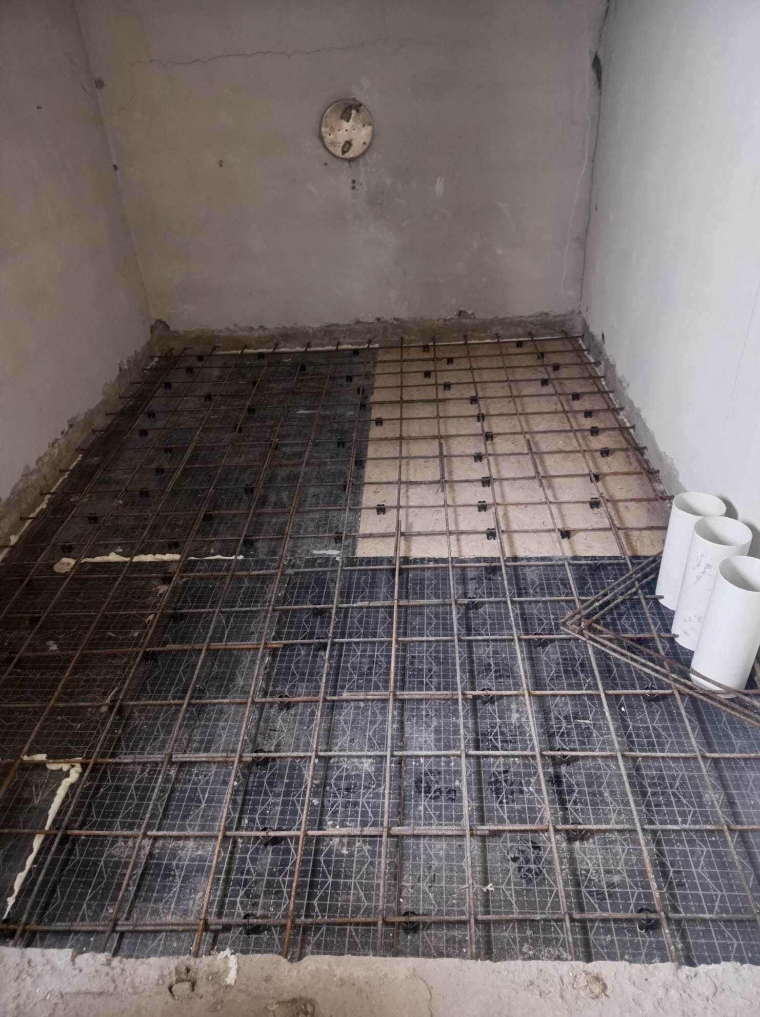 Przygotowanie zbrojenia pod wylewkę betonową w surowym wnętrzu, widoczne pręty zbrojeniowe ułożone na czarnej folii z dystansami, fragment podłoża bez folii, białe rury PCV w rogu pomieszczenia.
