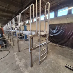 Konstrukcja pomostu roboczego ze stali nierdzewnej z drabinką, widoczna w hali produkcyjnej, surowy beton na podłodze, metalowe elementy czekają na montaż.