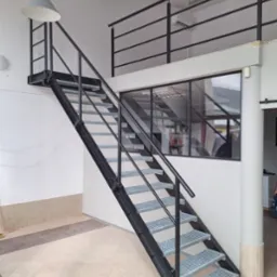 Nowoczesne schody z metalową konstrukcją i ażurowymi stopniami w Raciborzu, prowadzące na antresolę z czarną balustradą, oświetlone wiszącą lampą w industrialnym stylu.