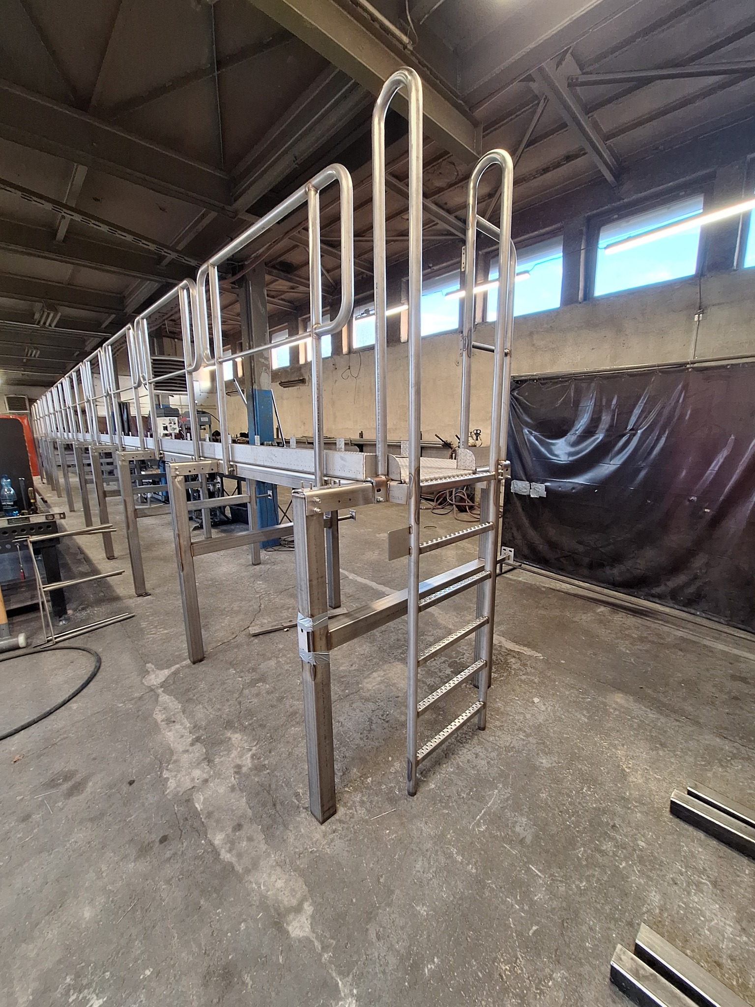 Konstrukcja pomostu roboczego ze stali nierdzewnej z drabinką, widoczna w hali produkcyjnej, surowy beton na podłodze, metalowe elementy czekają na montaż.