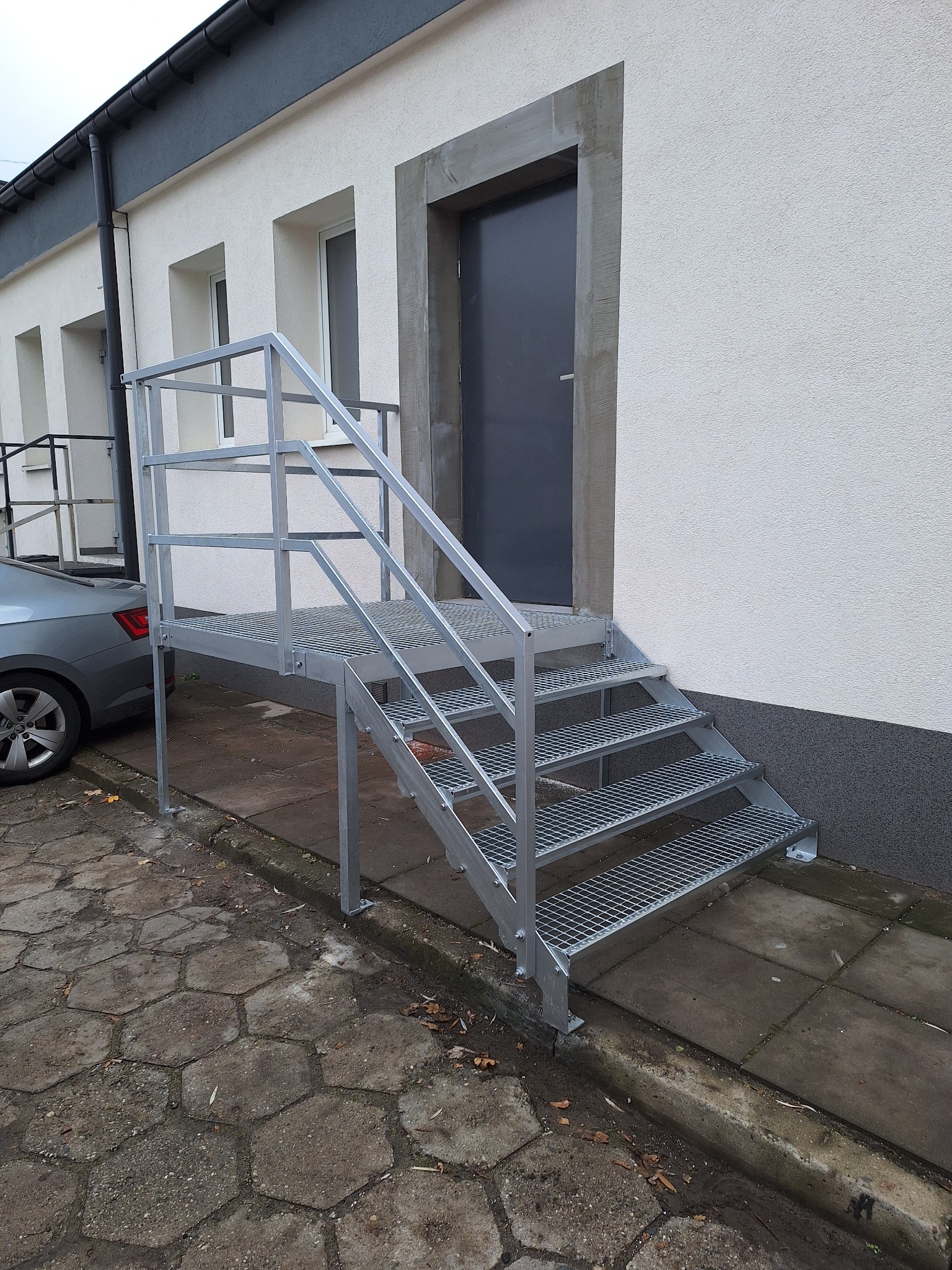 Zewnętrzne schody z metalową konstrukcją kratową prowadzące do drzwi budynku, widoczne z boku, z szarą elewacją i fragmentem zaparkowanego samochodu w tle.