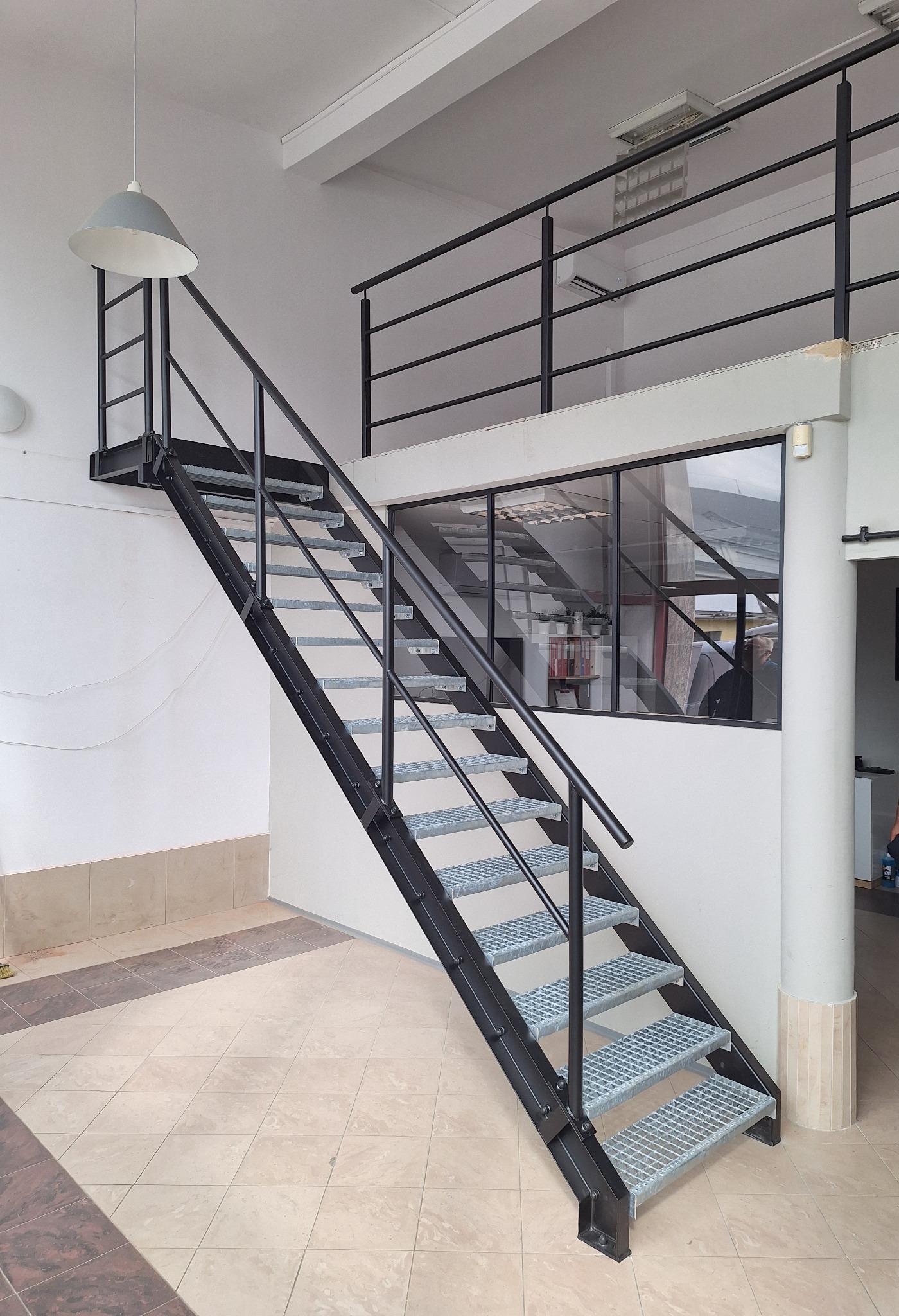 Nowoczesne schody z metalową konstrukcją i ażurowymi stopniami w Raciborzu, prowadzące na antresolę z czarną balustradą, oświetlone wiszącą lampą w industrialnym stylu.