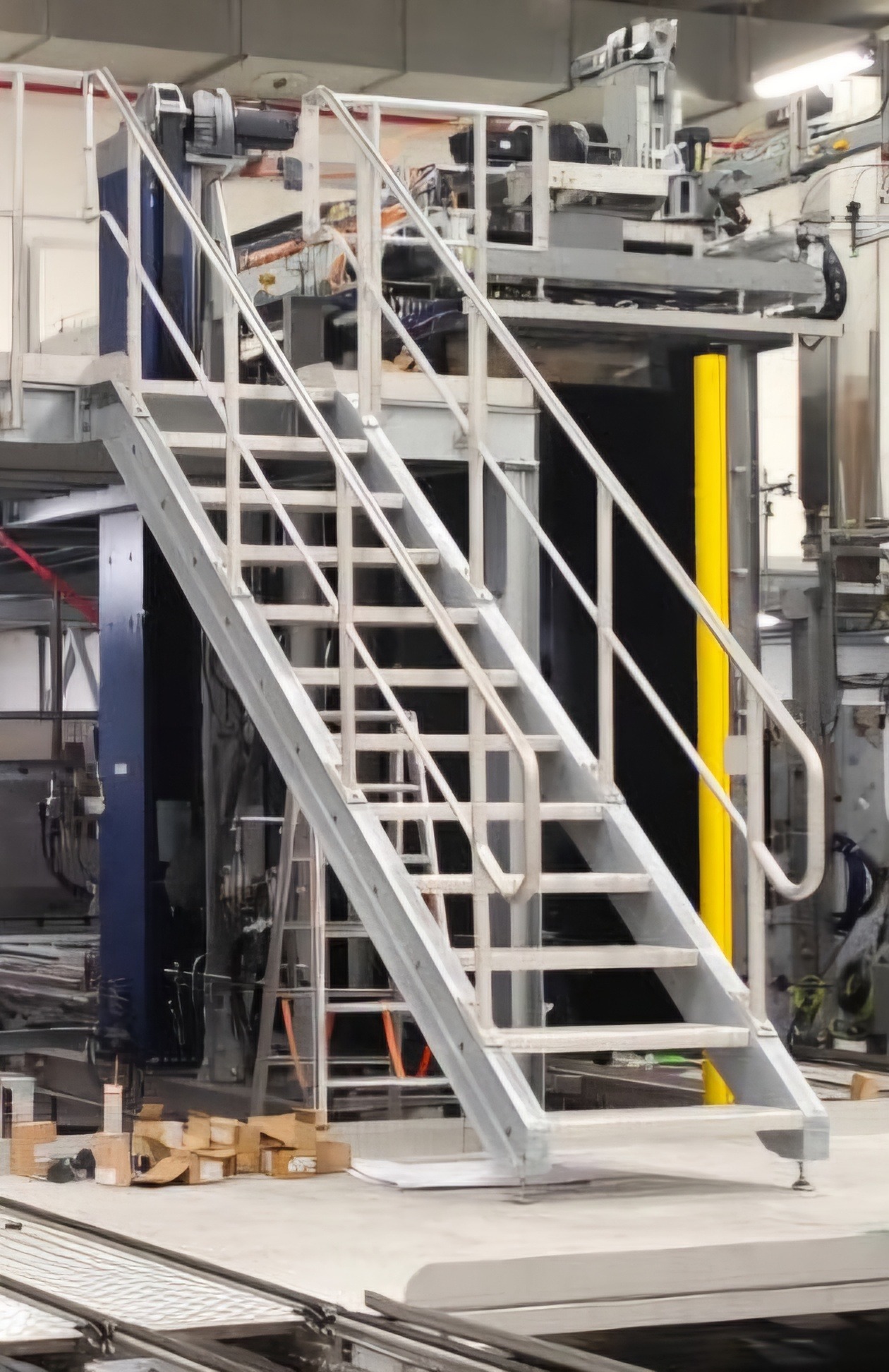 Szare, metalowe schody przemysłowe z barierkami, prowadzące do platformy z maszynami, w tle widoczne kartonowe pudełka i drabina.