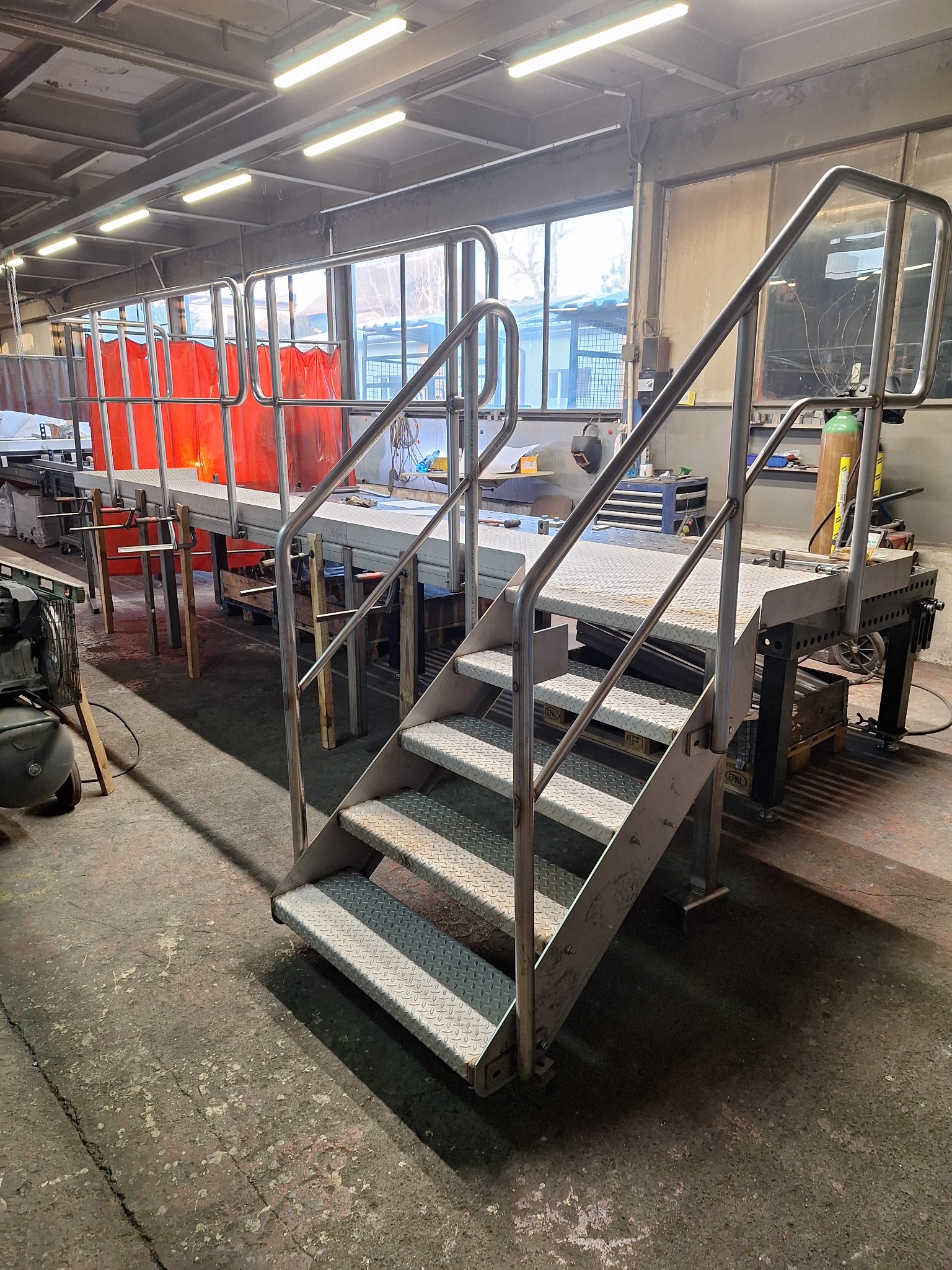 Metalowe schody z platformą roboczą w warsztacie, z widocznymi śladami użytkowania na podłodze i elementami wyposażenia w tle.