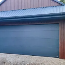 Szara segmentowa brama garażowa z poziomymi przetłoczeniami, zamontowana w brązowej drewnianej obudowie garażu z metalowym dachem, widok z zewnątrz na tle zieleni.