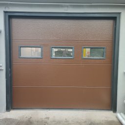 EIFAM Mateusz Nowacki - Brązowa segmentowa brama garażowa z trzema małymi okienkami, widoczna z zewnątrz, z szarą ramą i rurą spustową po lewej stronie, oraz materiałami budowlanymi po prawej stronie.