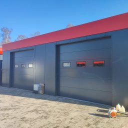 EIFAM Mateusz Nowacki - Dwa segmentowe wjazdy do hali przemysłowej w kolorze grafitowym z czerwonym pasem u góry, na kostce brukowej leży dmuchawa do liści i kanistry.