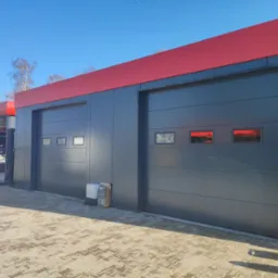 Dwie szare bramy segmentowe z czerwonymi nadprożami, widoczne okienka w bramach, na pierwszym planie kostka brukowa, obok bramy leżą narzędzia ogrodnicze.